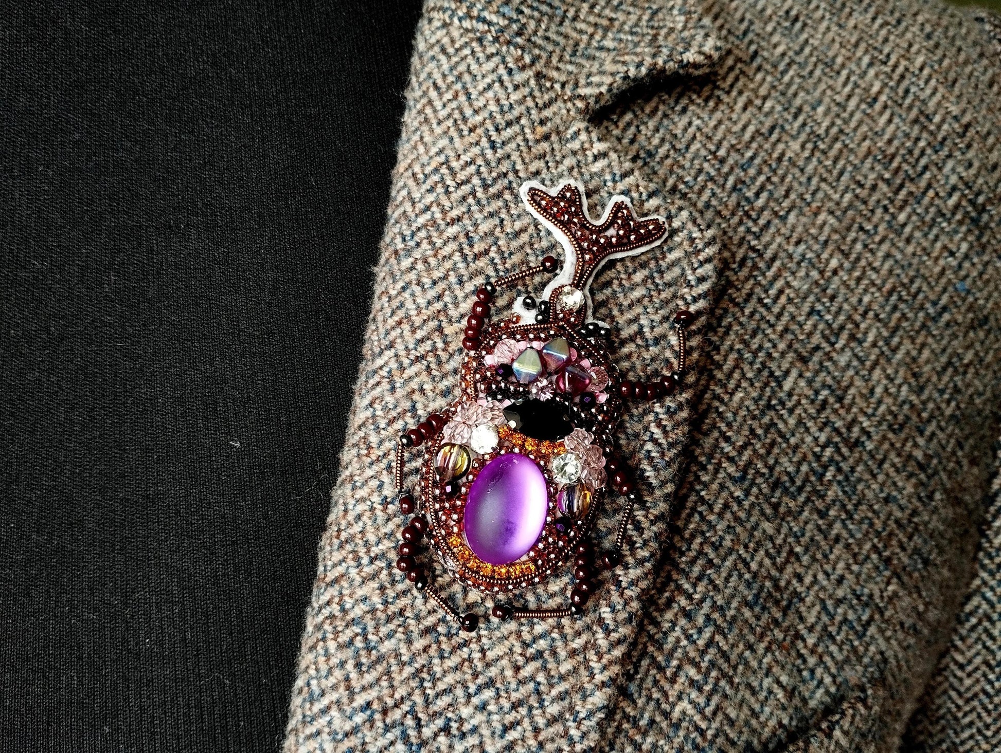 A close-up image of a decorative insect brooch with a purple gemstone, placed on a tweed jacket.