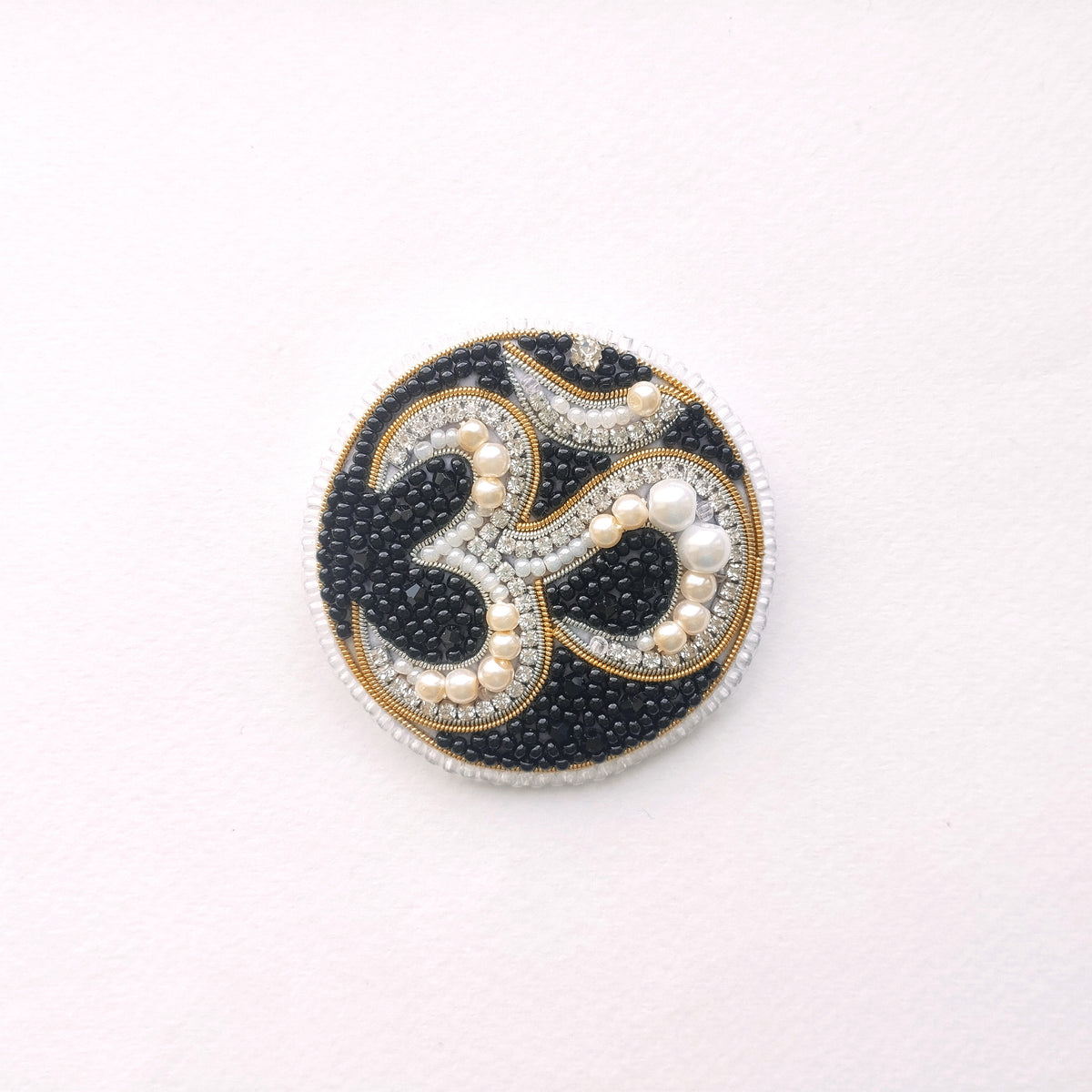 A circular, ornate piece of jewelry with a black and gold design, featuring the Om symbol and pearls.