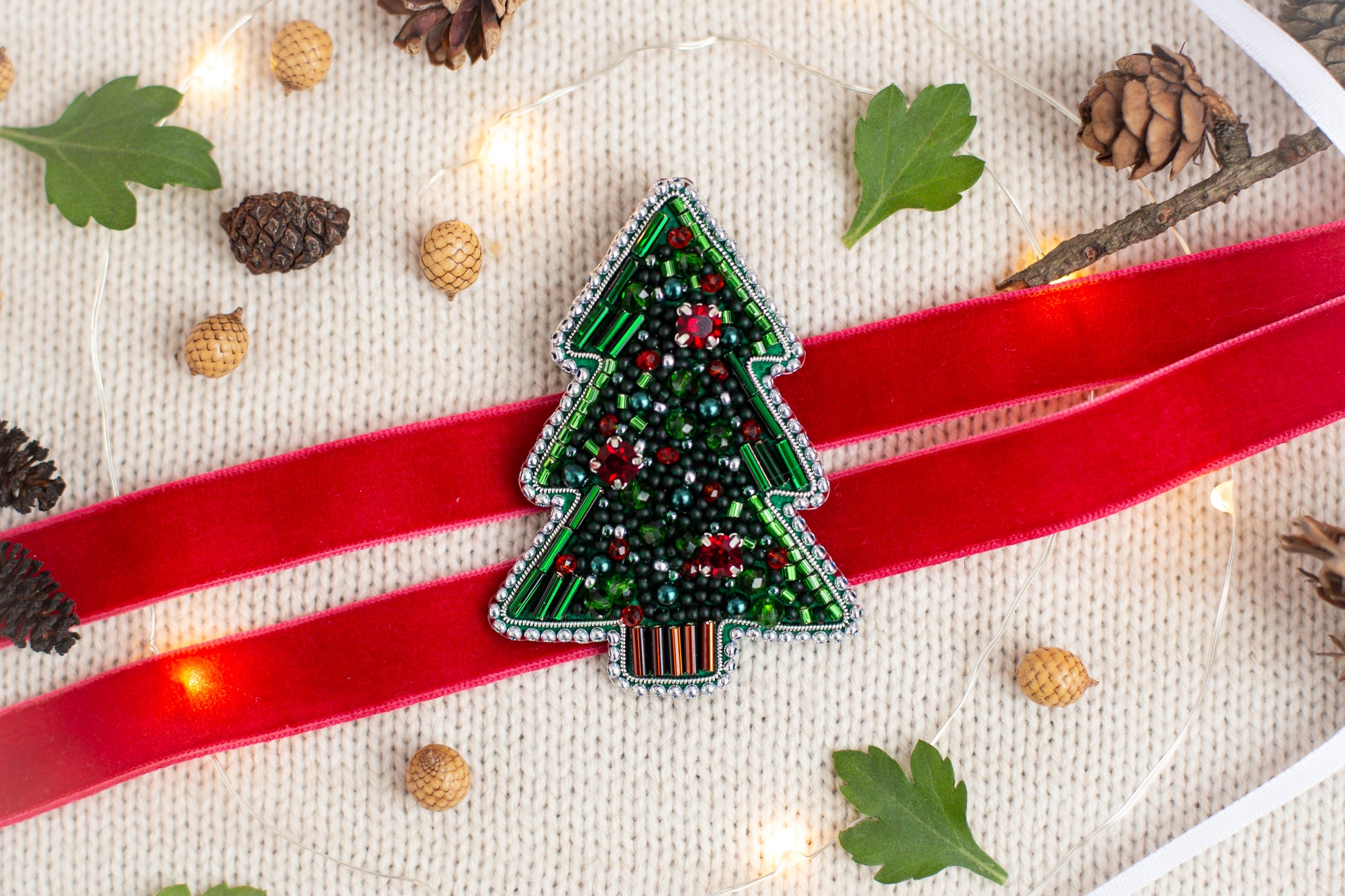 a Christmas tree-shaped ornament with red ribbon and beaded embellishments, surrounded by pine cones, a red ribbon, and twinkling lights.