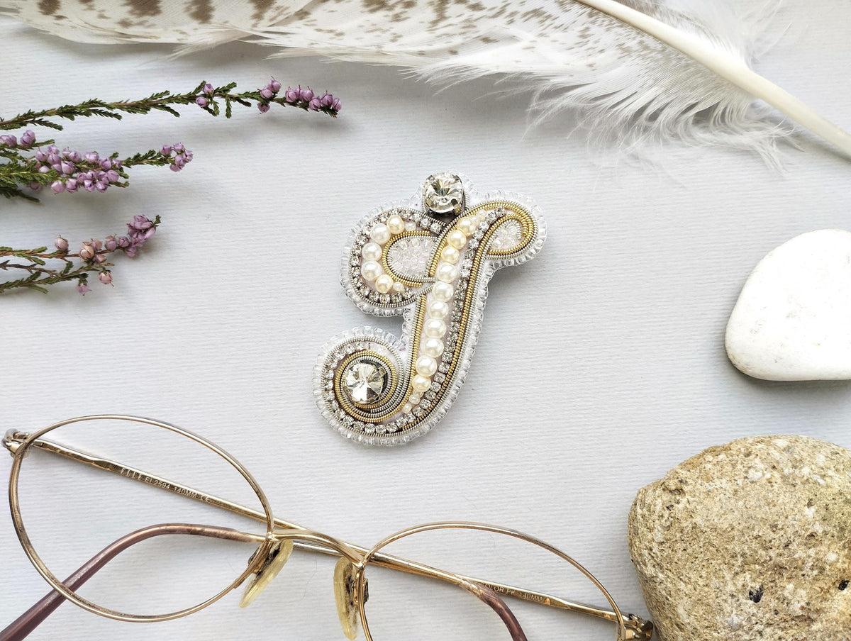 a close-up view of a decorative letter "J" made of beads and pearls, surrounded by various accessories such as glasses, feathers, and rocks.