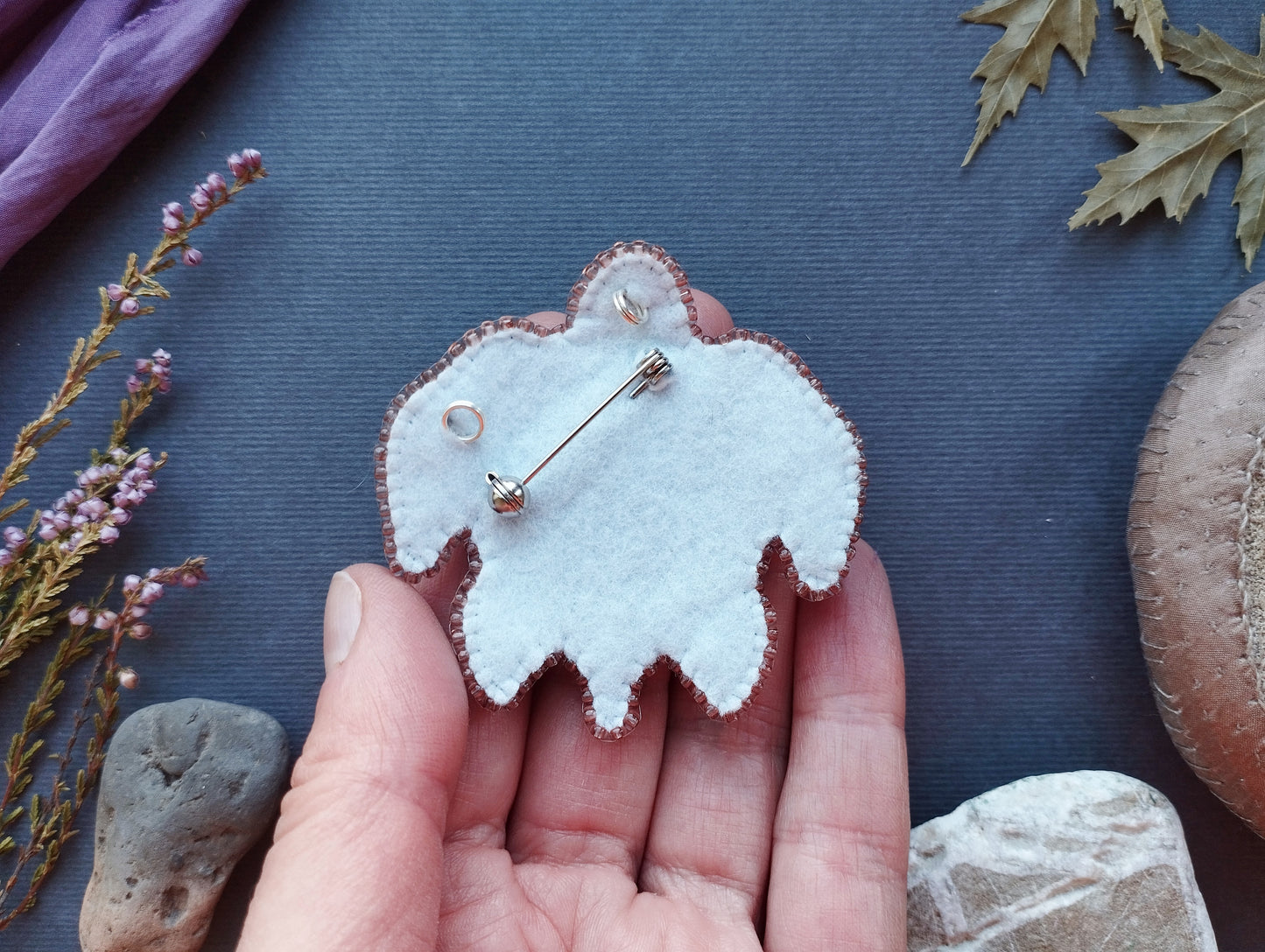 A hand holding a small, white felt object with a pin attached to it, surrounded by various natural elements such as leaves, rocks, and flowers.