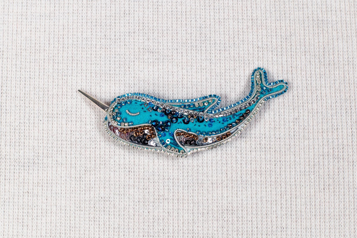 A decorative blue and silver brooch in the shape of a narwhal, with intricate beading and embellishments.