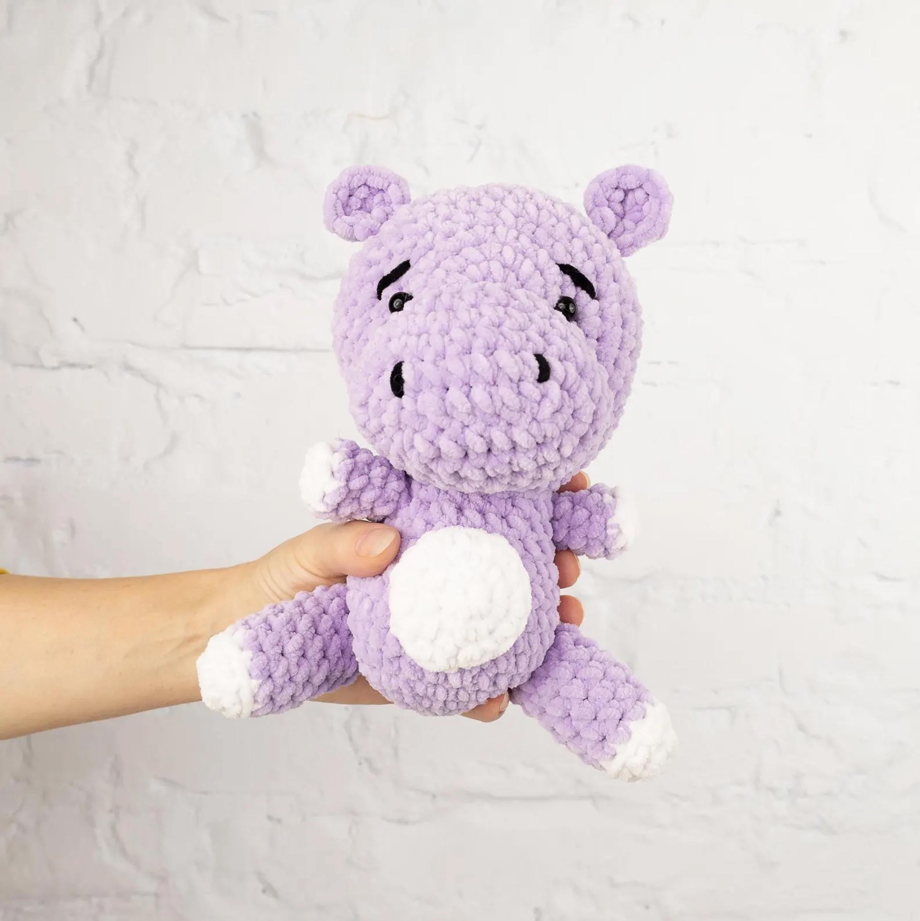 A hand is holding a purple crocheted stuffed animal, which appears to be a small, cute plush toy.