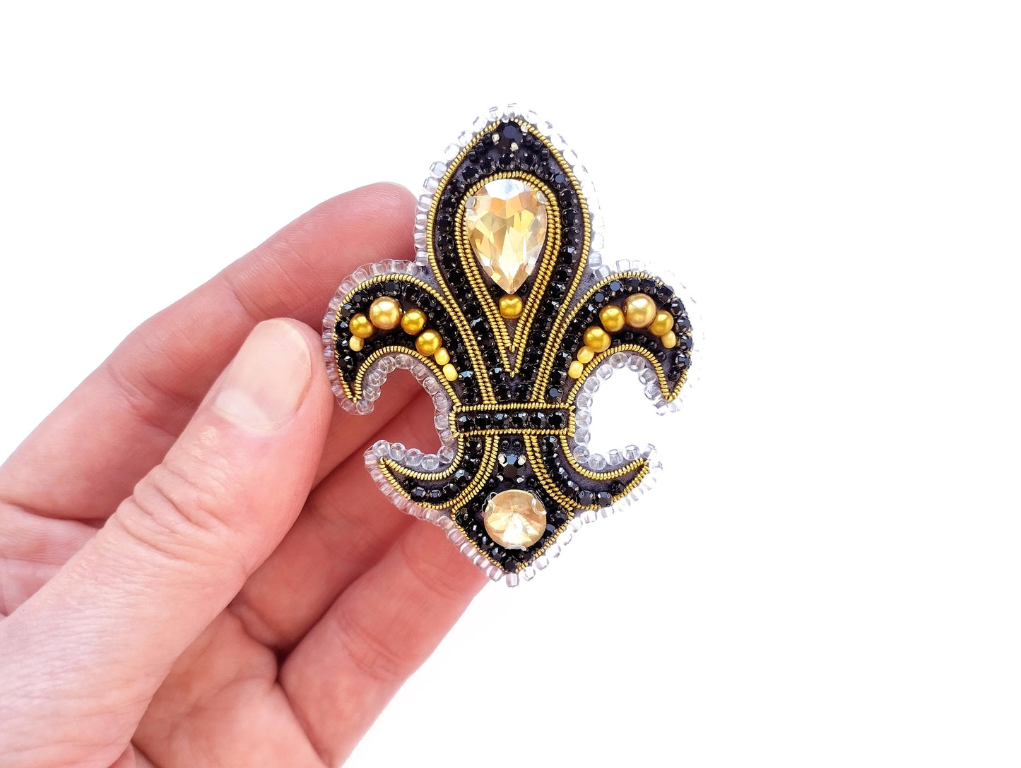 A hand holding a decorative brooch with a fleur-de-lis design, featuring a central yellow gem surrounded by smaller gold and black gemstones.