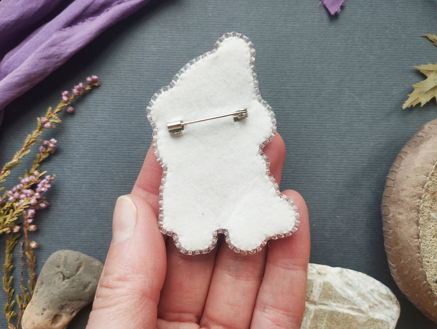 A hand holding a white felt object with a silver pin attached to it, surrounded by various natural elements such as leaves, rocks, and flowers.