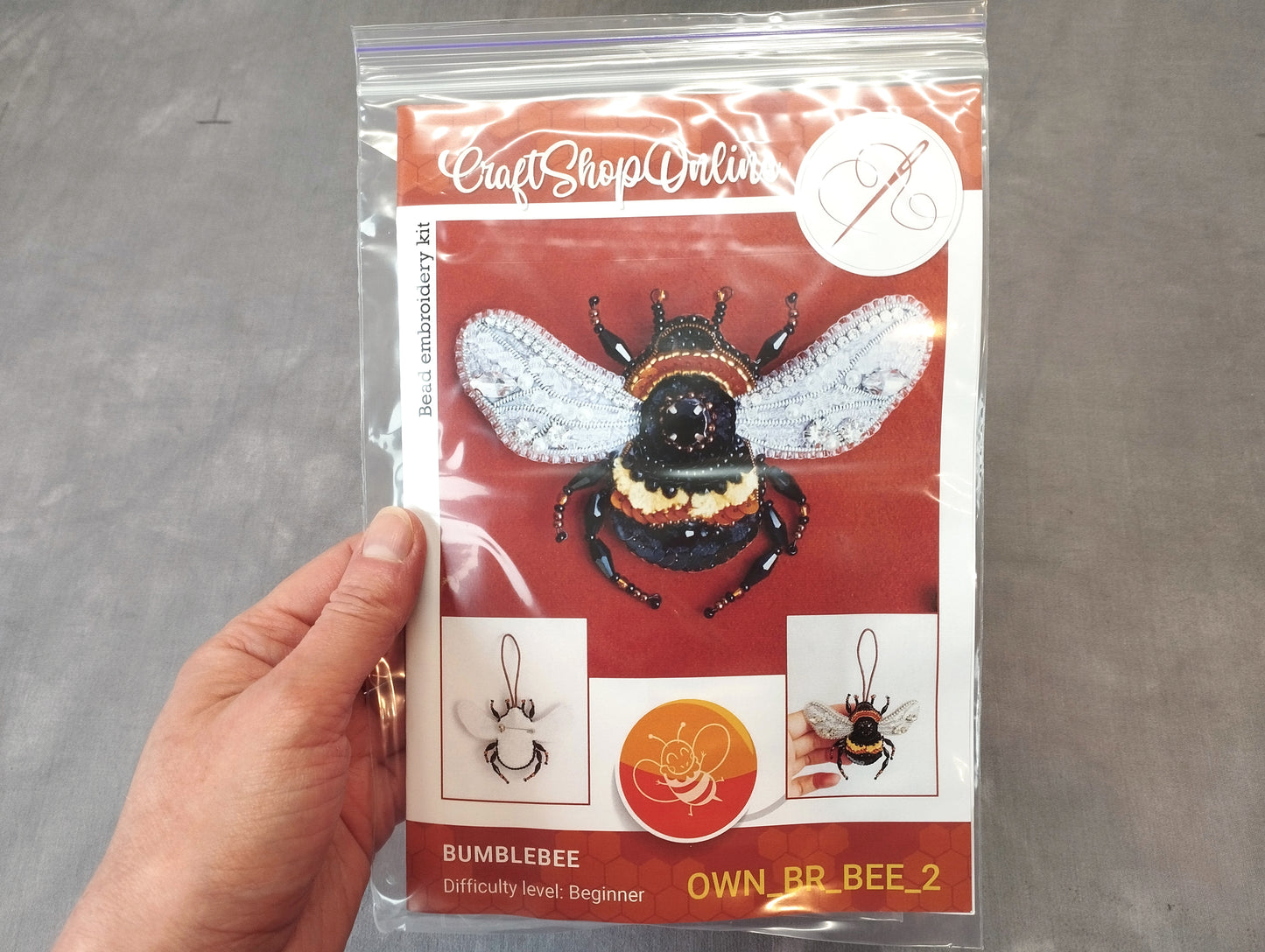 A hand holding a clear plastic bag containing a kit for creating a bumblebee embroidery piece.