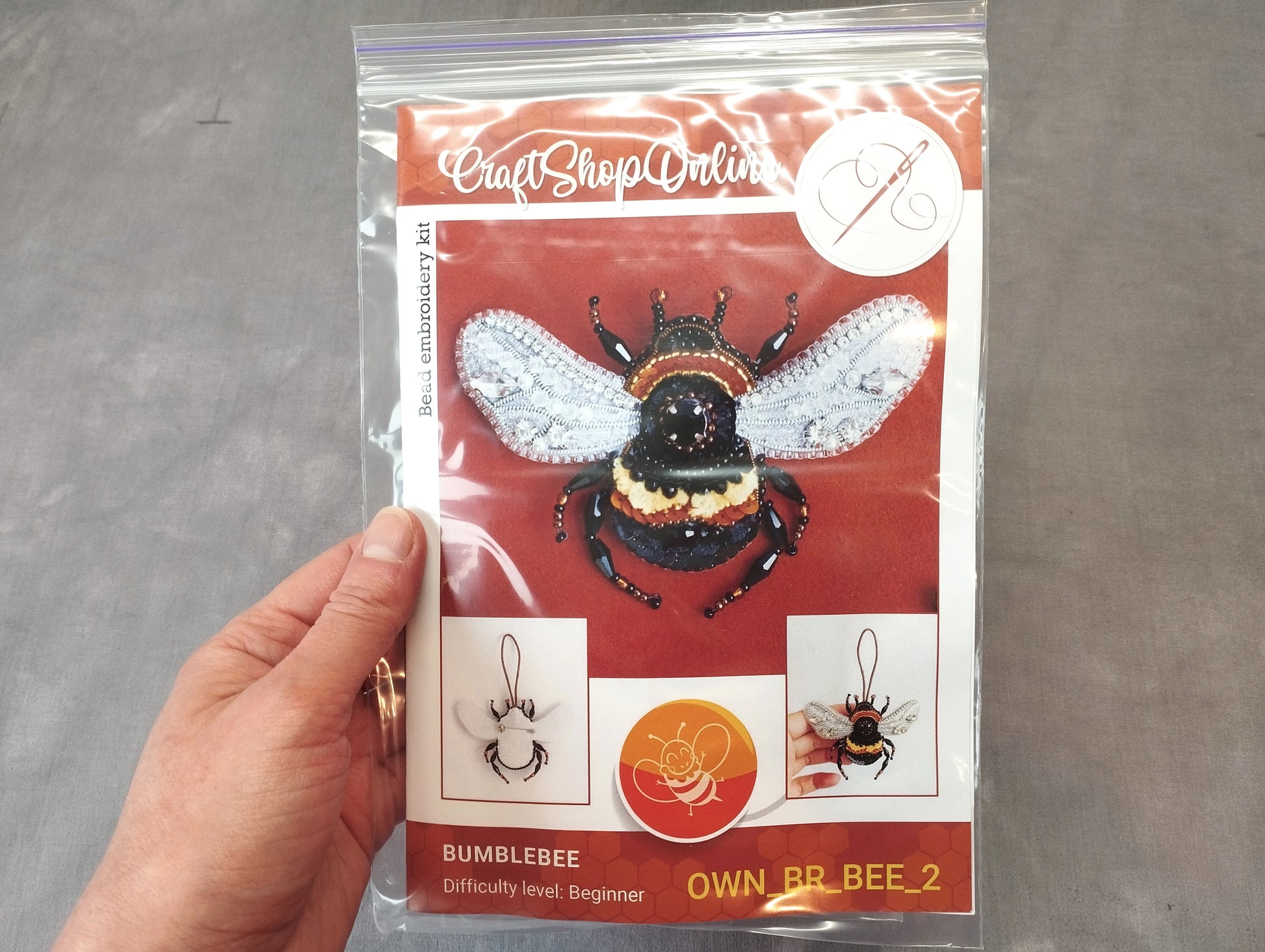 A hand holding a clear plastic bag containing a kit for creating a bumblebee embroidery piece.