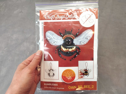 A hand holding a clear plastic bag containing a kit for creating a bumblebee embroidery piece.