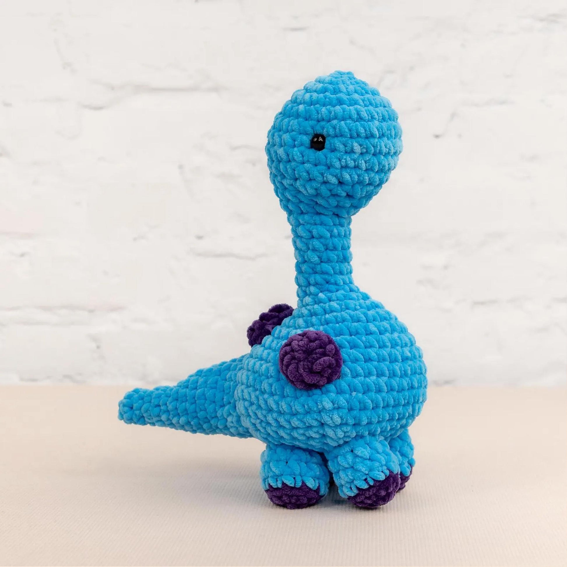 A blue crocheted toy dinosaur with purple accents is sitting on a white surface against a white brick wall background.