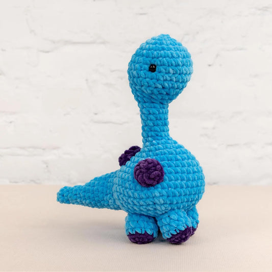 A blue crocheted toy dinosaur with purple accents is sitting on a white surface against a white brick wall background.