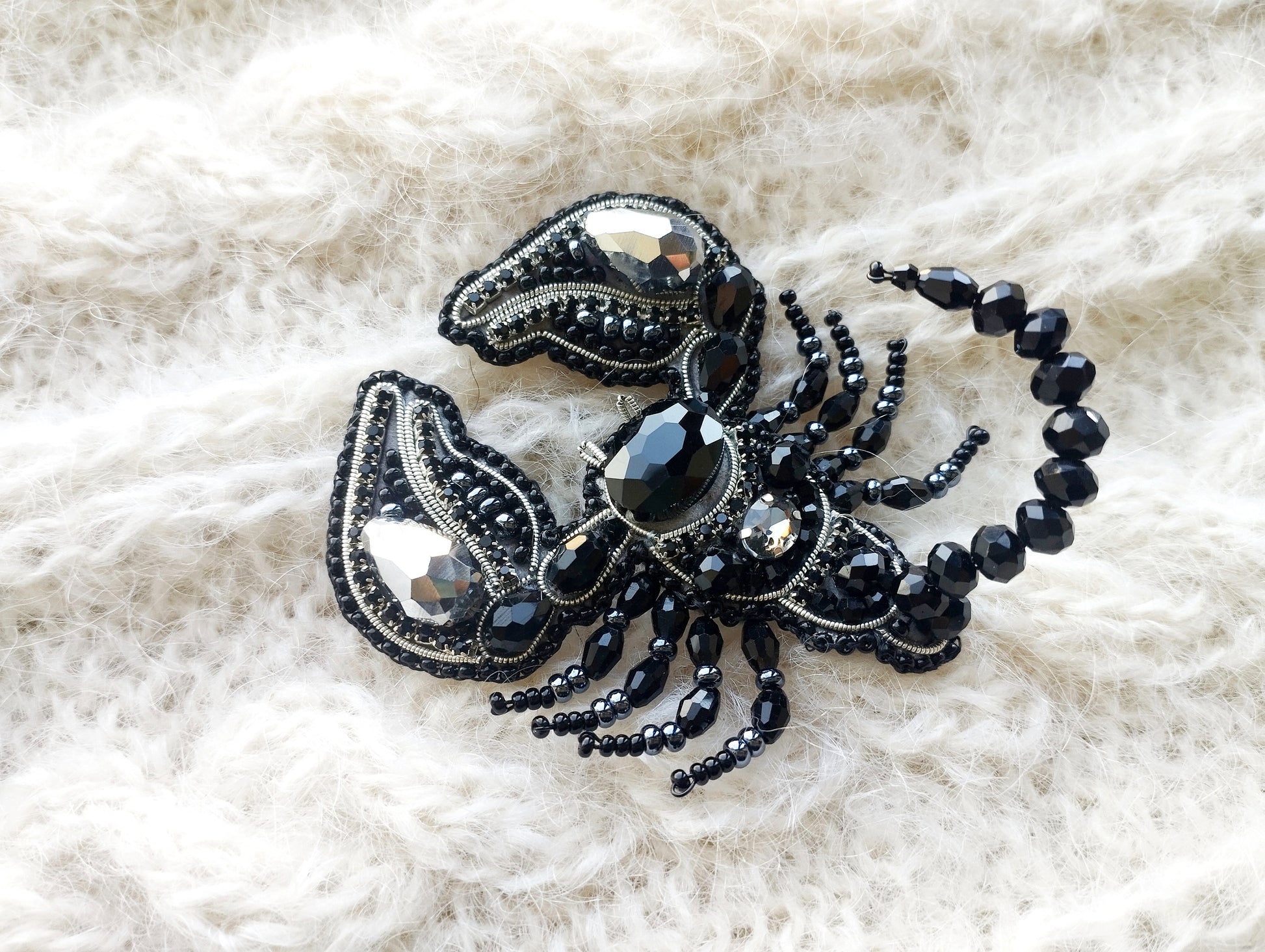 A close-up image of a decorative black and silver piece of jewelry, possibly a brooch or pin, resting on a white, textured fabric background.