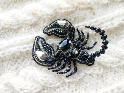 A close-up image of a decorative black and silver piece of jewelry, possibly a brooch or pin, resting on a white, textured fabric background.