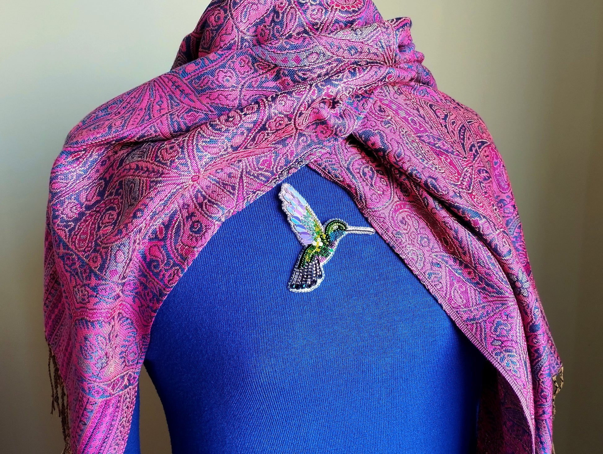 A mannequin wearing a blue top and a pink and purple paisley-patterned shawl. The shawl is draped over the mannequin's shoulders, and there is a small, colorful bird brooch pinned to the mannequin's shirt.
