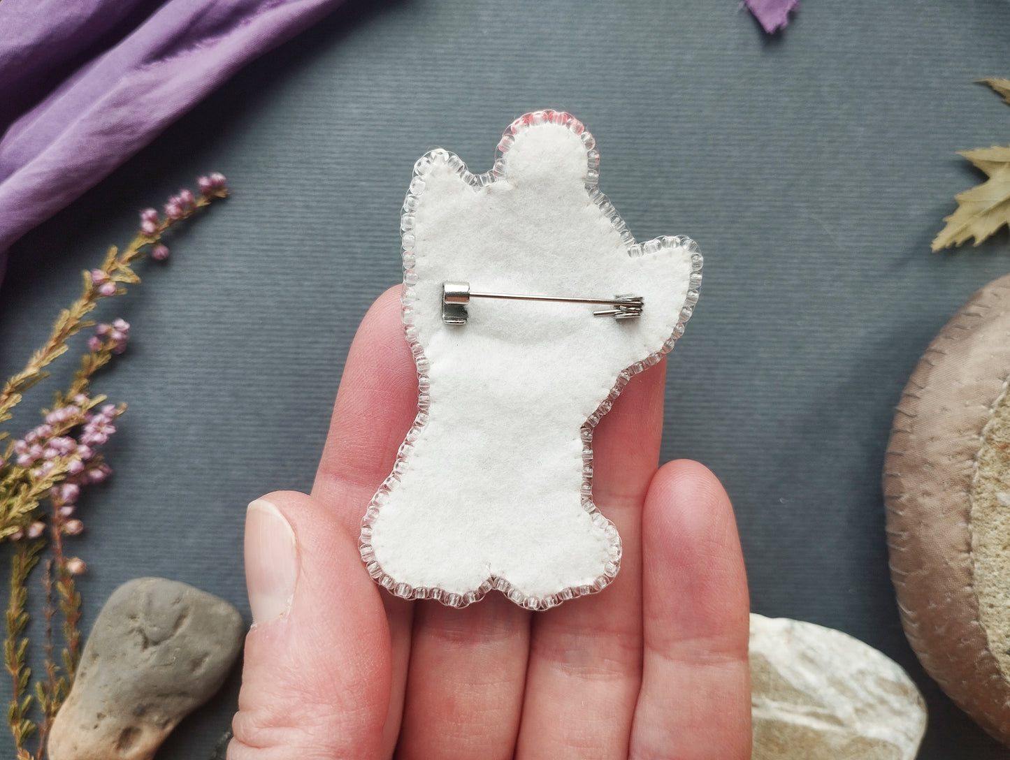 A hand holding a white felt cat-shaped pin against a dark background with various natural elements, such as leaves and flowers.