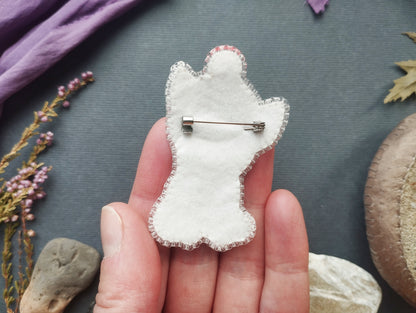 A hand holding a white felt cat-shaped pin against a dark background with various natural elements, such as leaves and flowers.