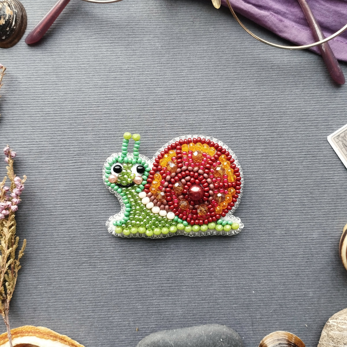 A colorful beaded snail-shaped object is displayed on a gray surface, surrounded by various objects such as a pair of glasses, a plant, and a small rock.