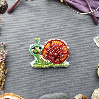 A colorful beaded snail-shaped object is displayed on a gray surface, surrounded by various objects such as a pair of glasses, a plant, and a small rock.