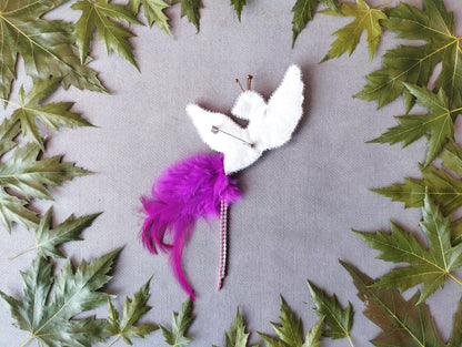 a white bird-shaped object with a purple feather attached to it, surrounded by green leaves on a gray background.