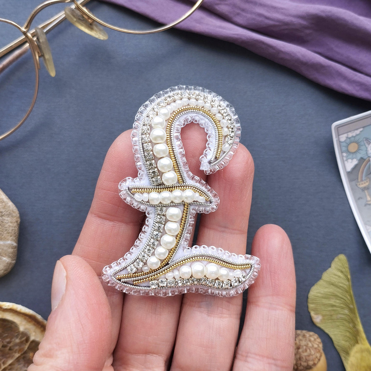 A hand holding a decorative monogram made of pearls and beads, with a pair of glasses and a purple fabric in the background.