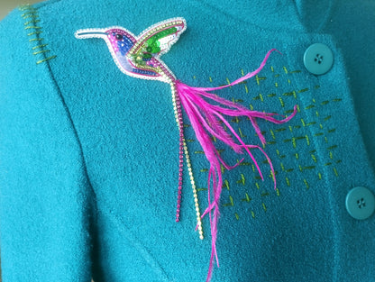 A close-up image of a teal-colored jacket with a decorative bird brooch featuring a colorful hummingbird design and a long pink feather.