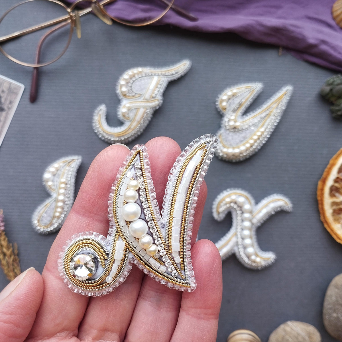 a hand holding a decorative letter n made of beads and pearls, with other decorative elements such as pearls and a pair of glasses in the background.