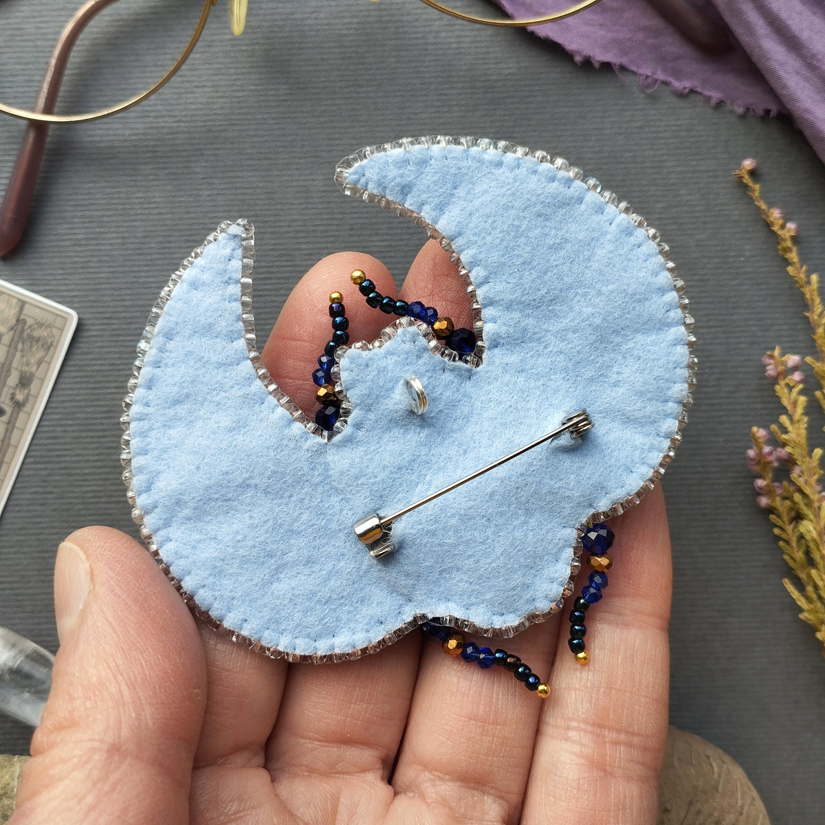 A hand holding a light blue felt crescent moon pin with blue and gold beads attached to it.