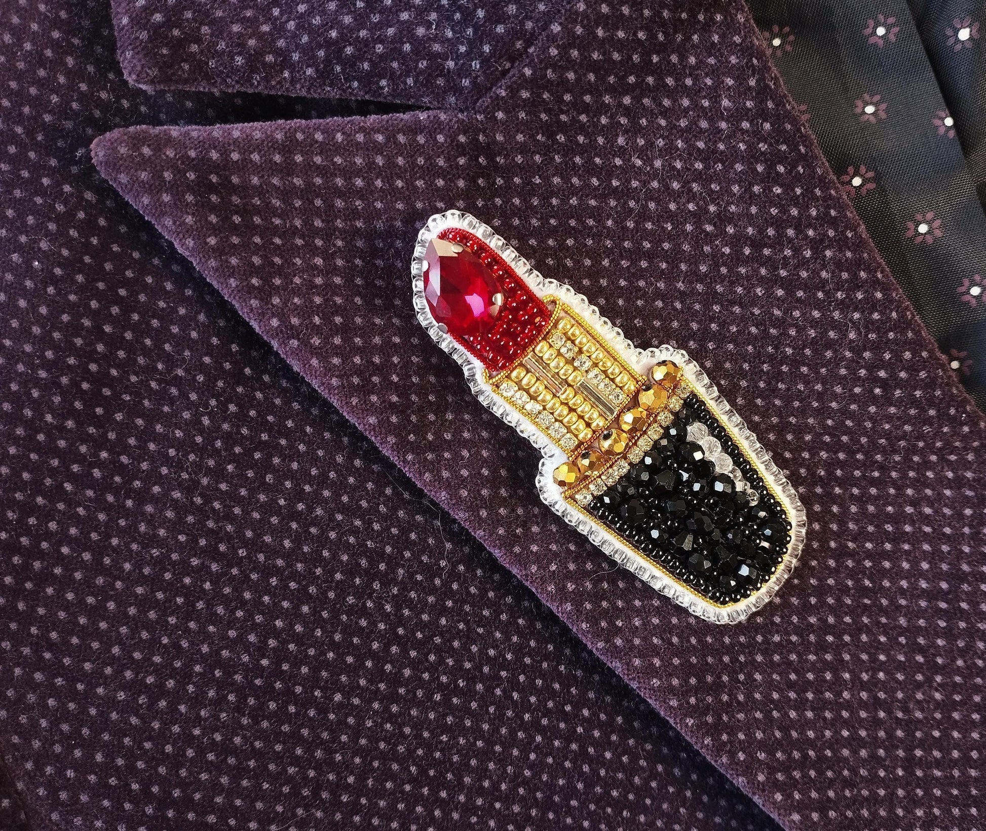A close-up image of a purple fabric with a decorative red and gold pin featuring a red lipstick design.