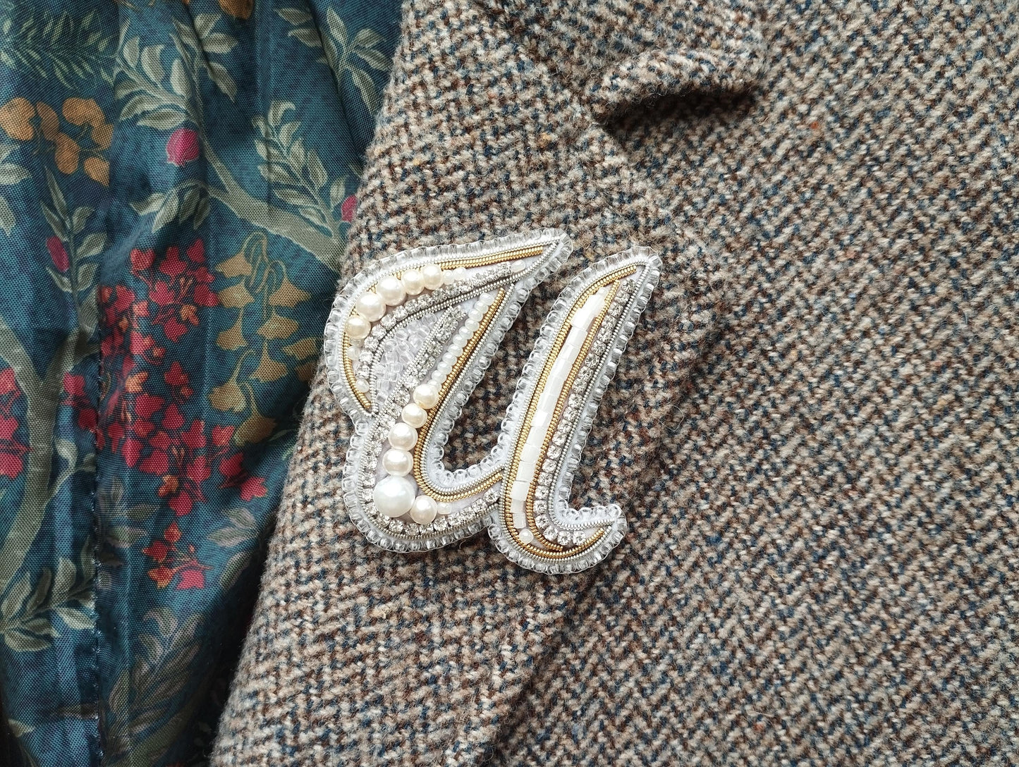 a close-up view of a decorative brooch or pin with pearls and a metallic design, attached to a tweed jacket or blazer.