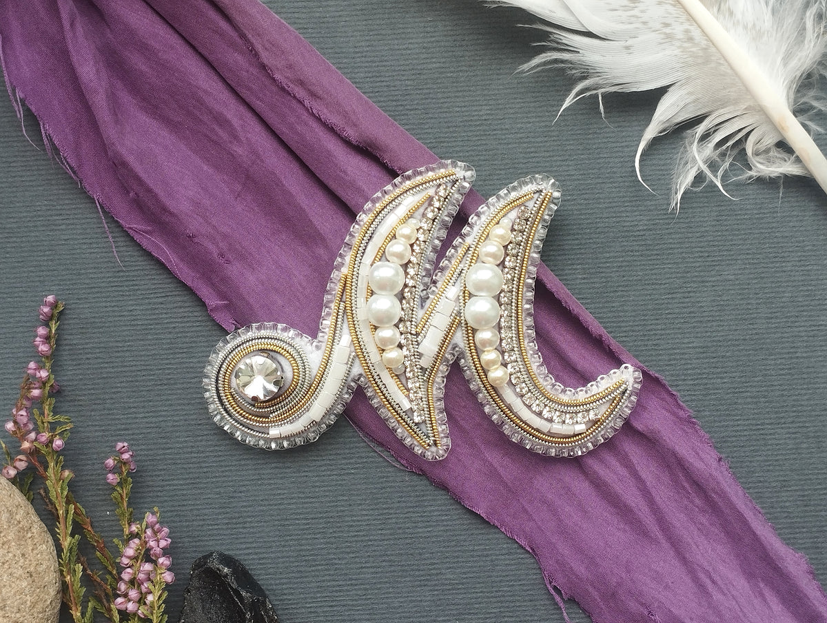 a decorative monogrammed letter "M" with a crystal embellishment, placed on a purple fabric. Surrounding the letter are various decorative elements, including a feather and some flowers, creating a visually appealing and artistic composition.