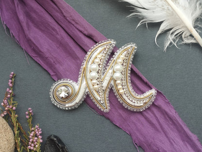 a decorative monogrammed letter "M" with a crystal embellishment, placed on a purple fabric. Surrounding the letter are various decorative elements, including a feather and some flowers, creating a visually appealing and artistic composition.