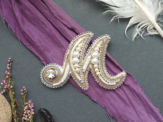 a decorative monogrammed letter "M" with a crystal embellishment, placed on a purple fabric. Surrounding the letter are various decorative elements, including a feather and some flowers, creating a visually appealing and artistic composition.