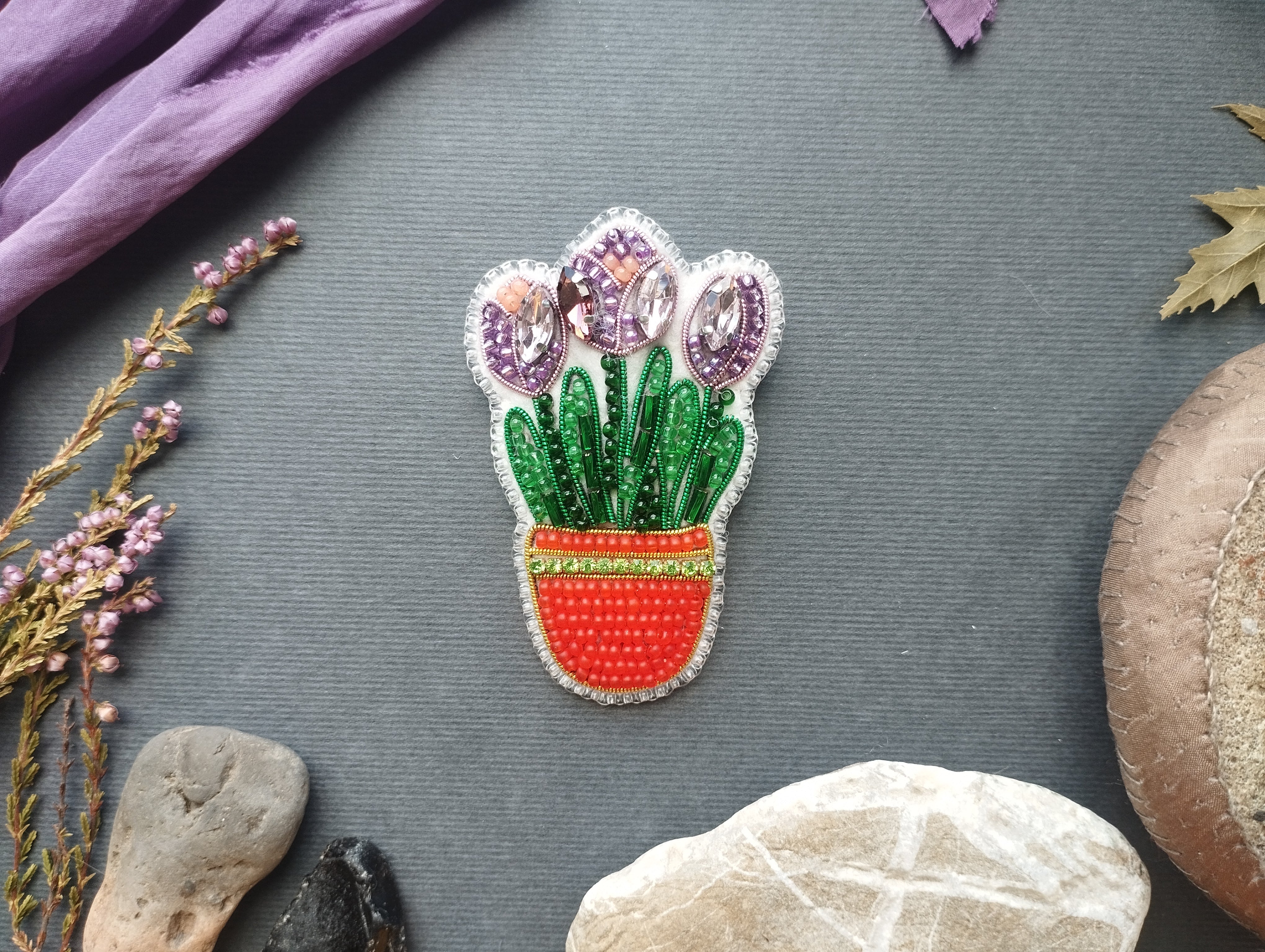 a decorative piece of embroidered flowerwork with purple and green elements, placed on a gray background. Surrounding the flowerwork are various natural elements, including a purple fabric, dried leaves, and rocks.