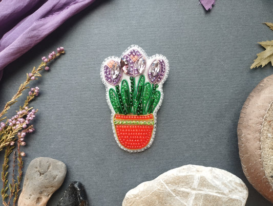 a decorative piece of embroidered flowerwork with purple and green elements, placed on a gray background. Surrounding the flowerwork are various natural elements, including a purple fabric, dried leaves, and rocks.