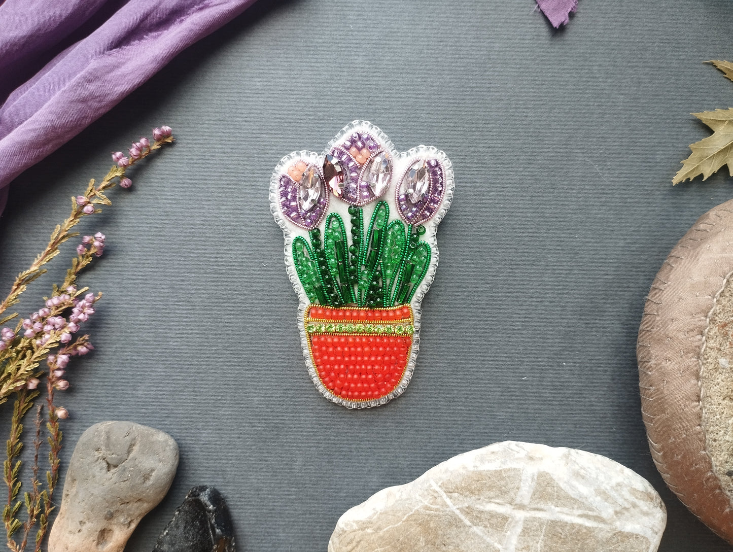 a decorative piece of embroidered flowerwork with purple and green elements, placed on a gray background. Surrounding the flowerwork are various natural elements, including a purple fabric, dried leaves, and rocks.