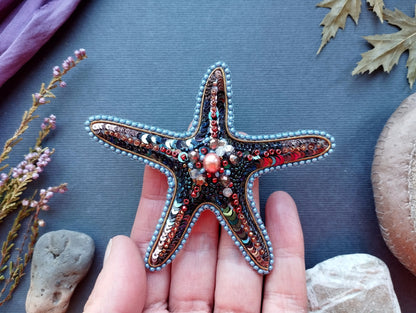 A hand holding a starfish adorned with colorful beads and sequins, surrounded by various natural elements such as leaves, rocks, and flowers.