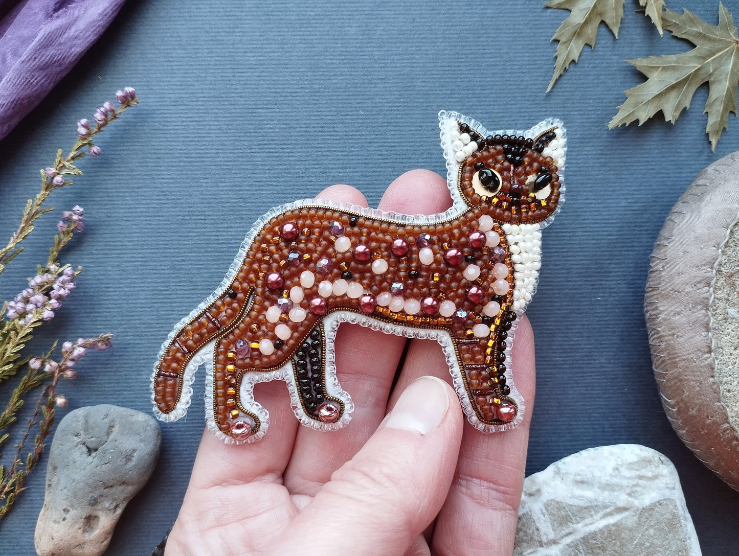A hand is holding a beaded embroidered cat pin against a blue background, surrounded by various natural elements such as leaves, rocks, and flowers.