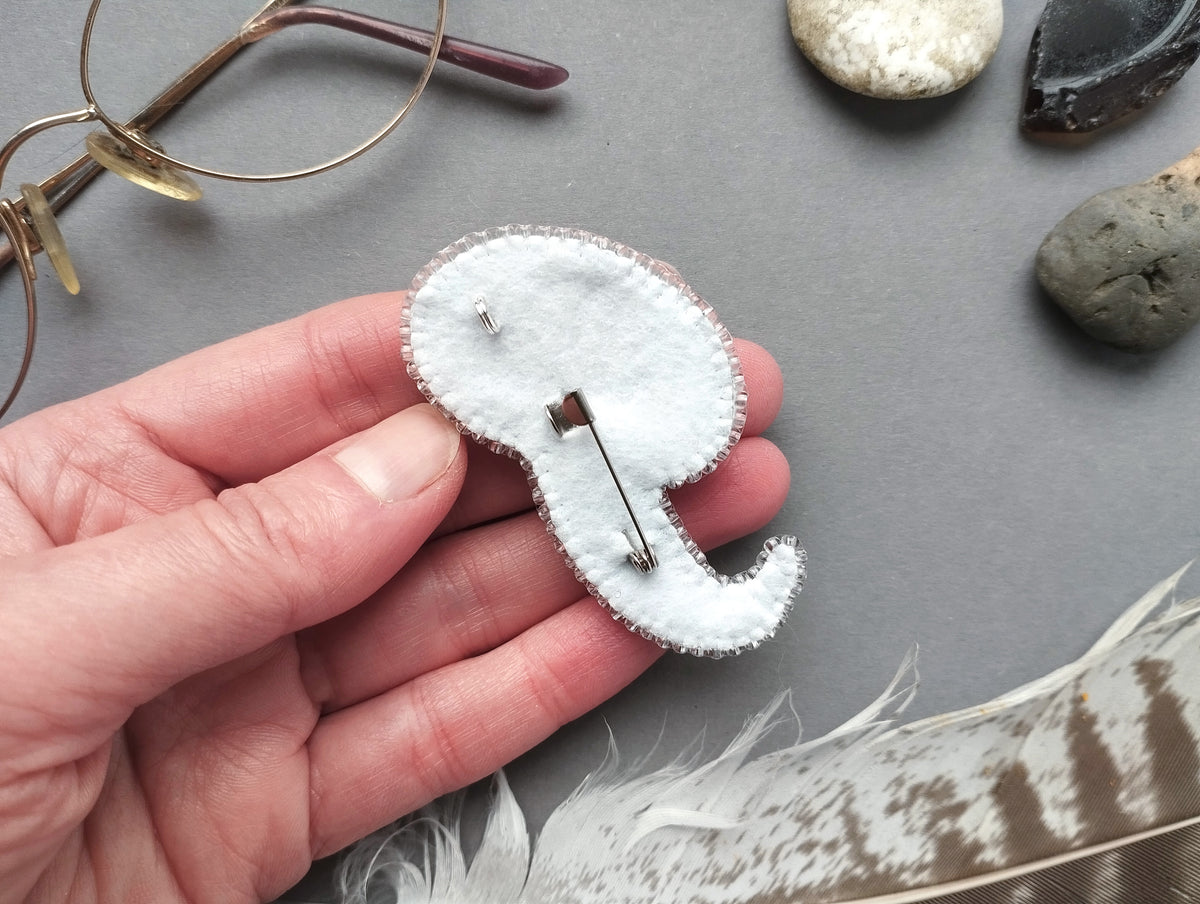 A hand holding a small, white felt object resembling a pin or pin cushion, surrounded by various objects such as glasses, rocks, and feathers.