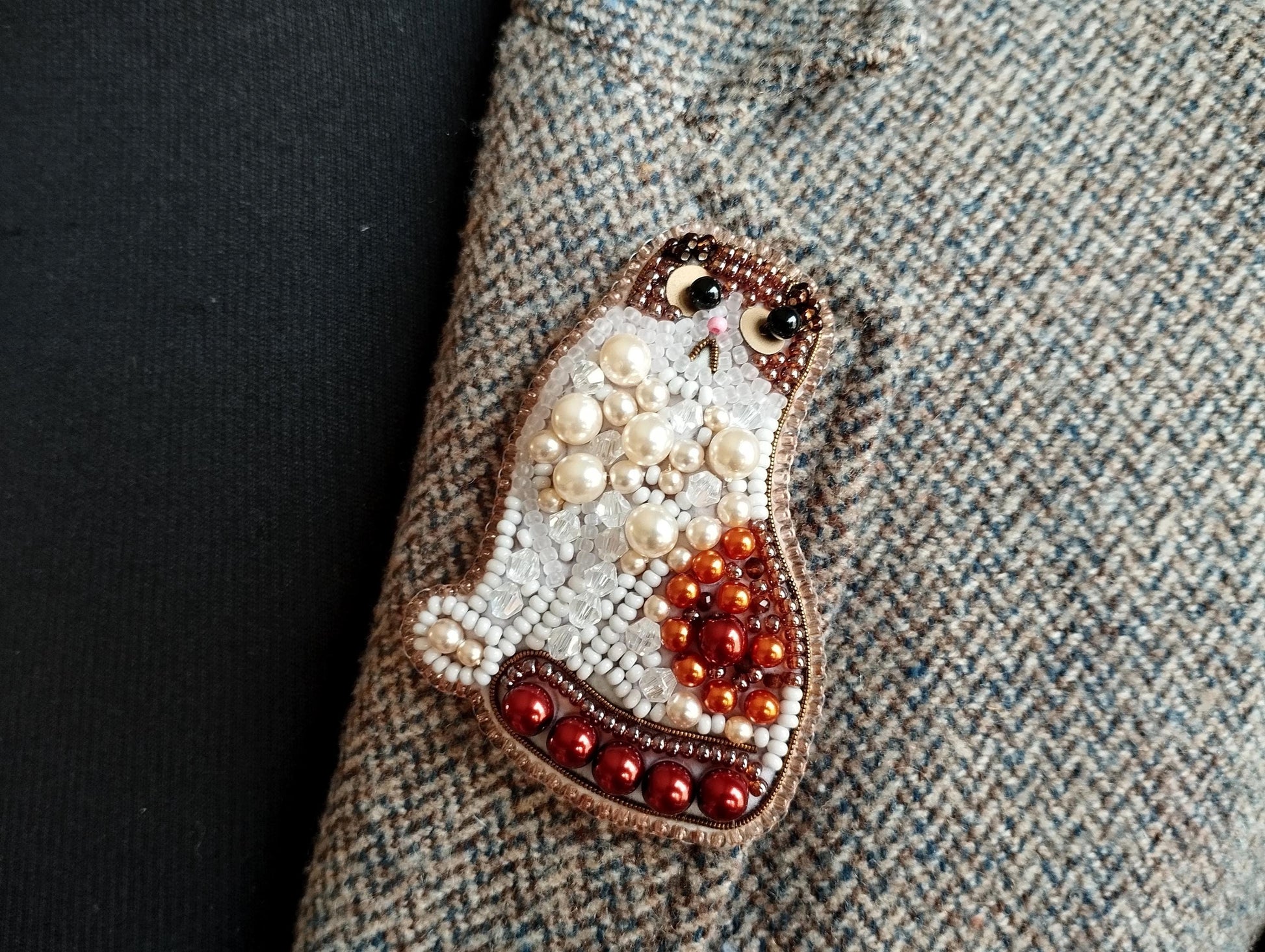 A close-up image of a decorative cat pin made of beads and pearls, placed on a textured fabric surface.