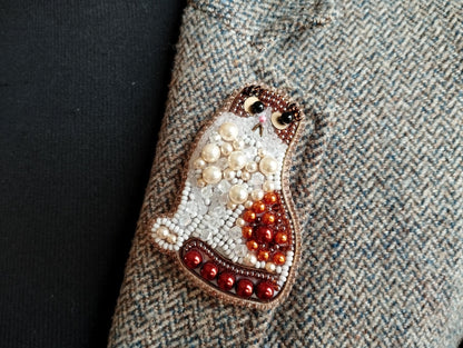 A close-up image of a decorative cat pin made of beads and pearls, placed on a textured fabric surface.