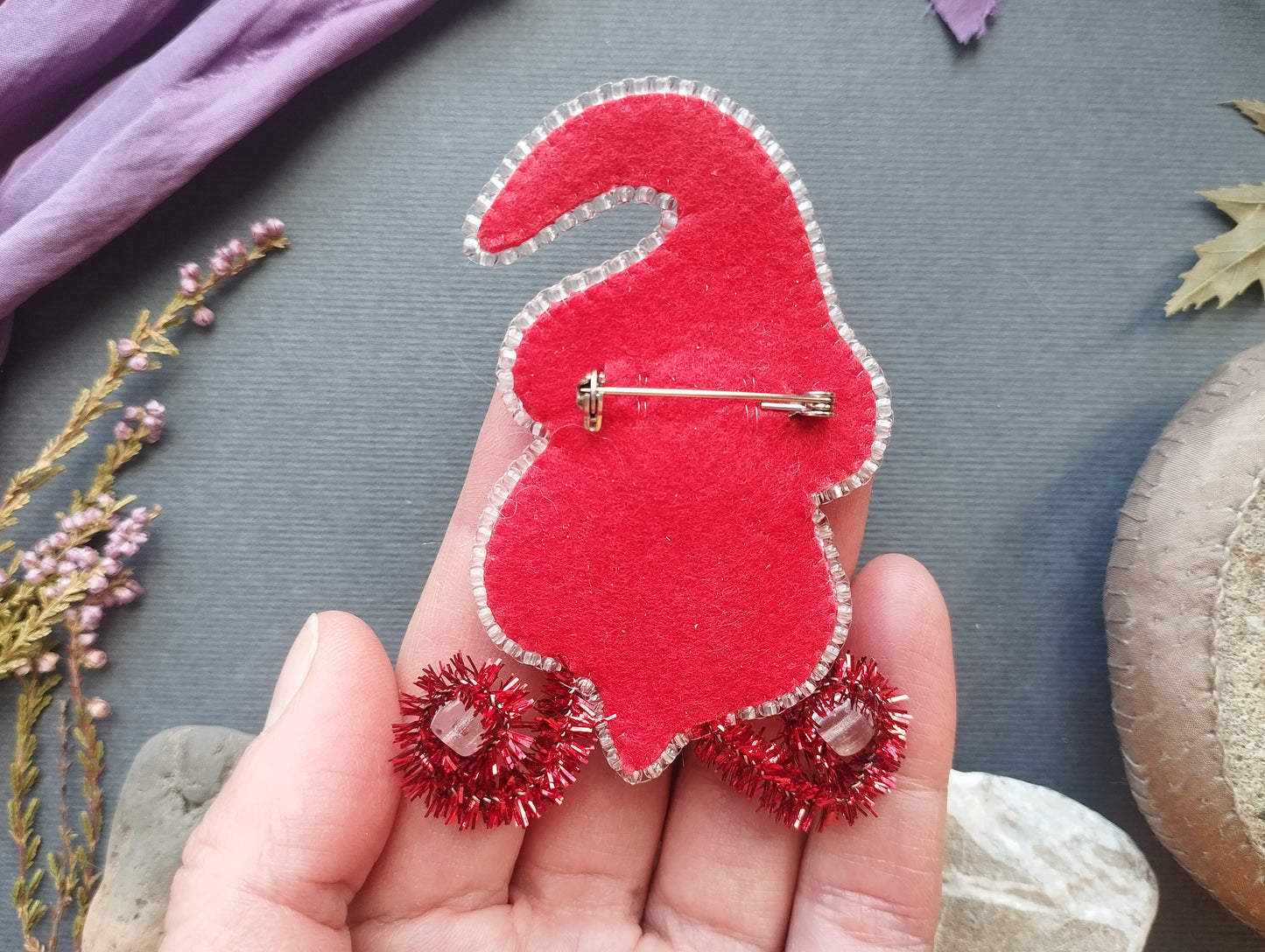 A hand holding a red felt gnome pin with red tinsel on its feet, surrounded by various decorative items such as flowers and leaves.
