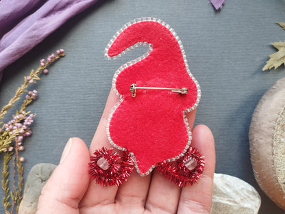 A hand holding a red felt gnome pin with red tinsel on its feet, surrounded by various decorative items such as flowers and leaves.