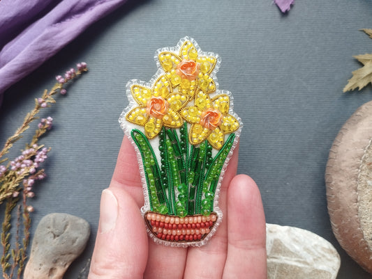 A hand holding a decorative beaded piece featuring a vase with yellow flowers and green leaves.