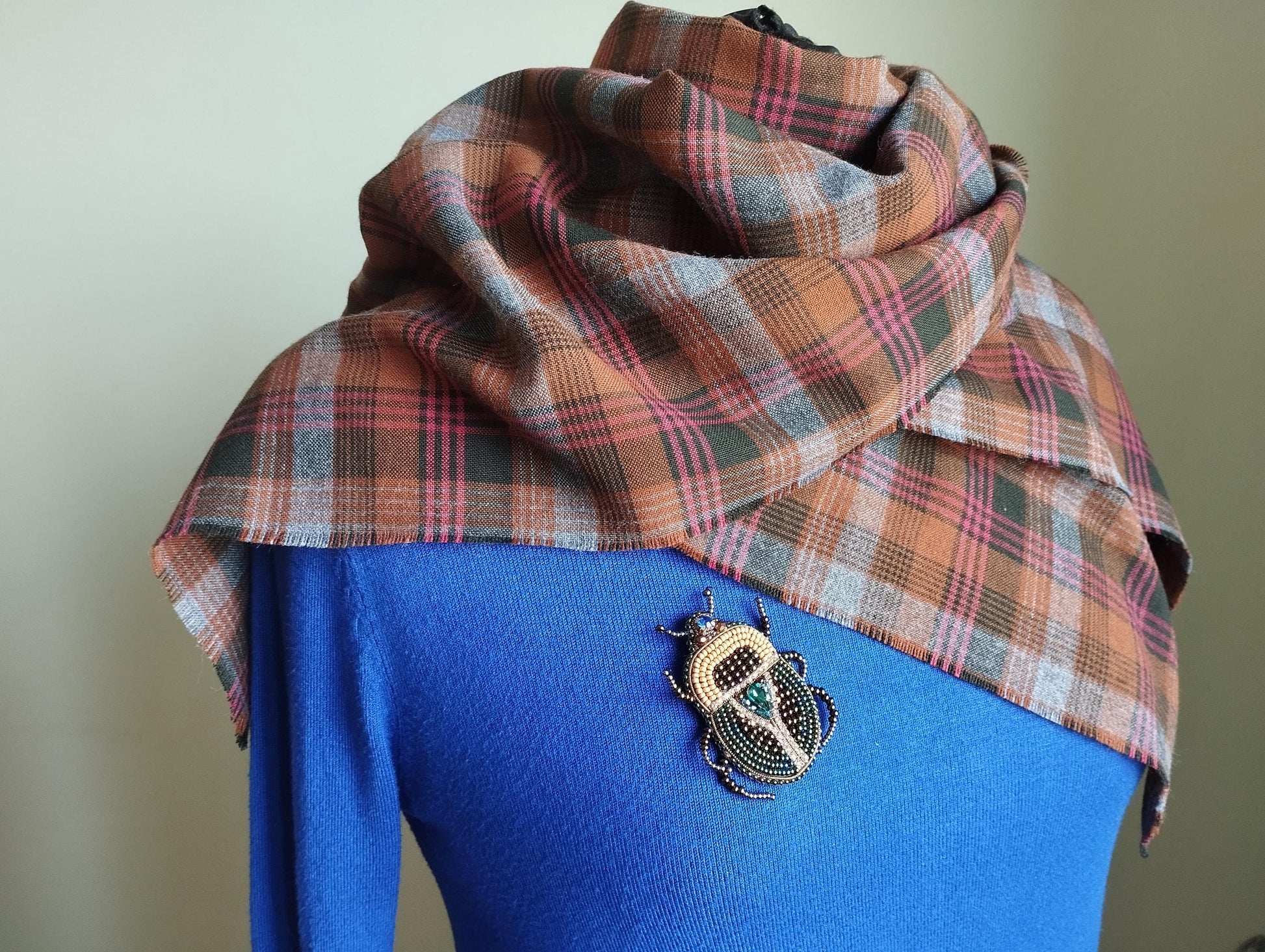 A plaid scarf is draped over a blue sweater, with a decorative insect brooch pinned to the sweater.