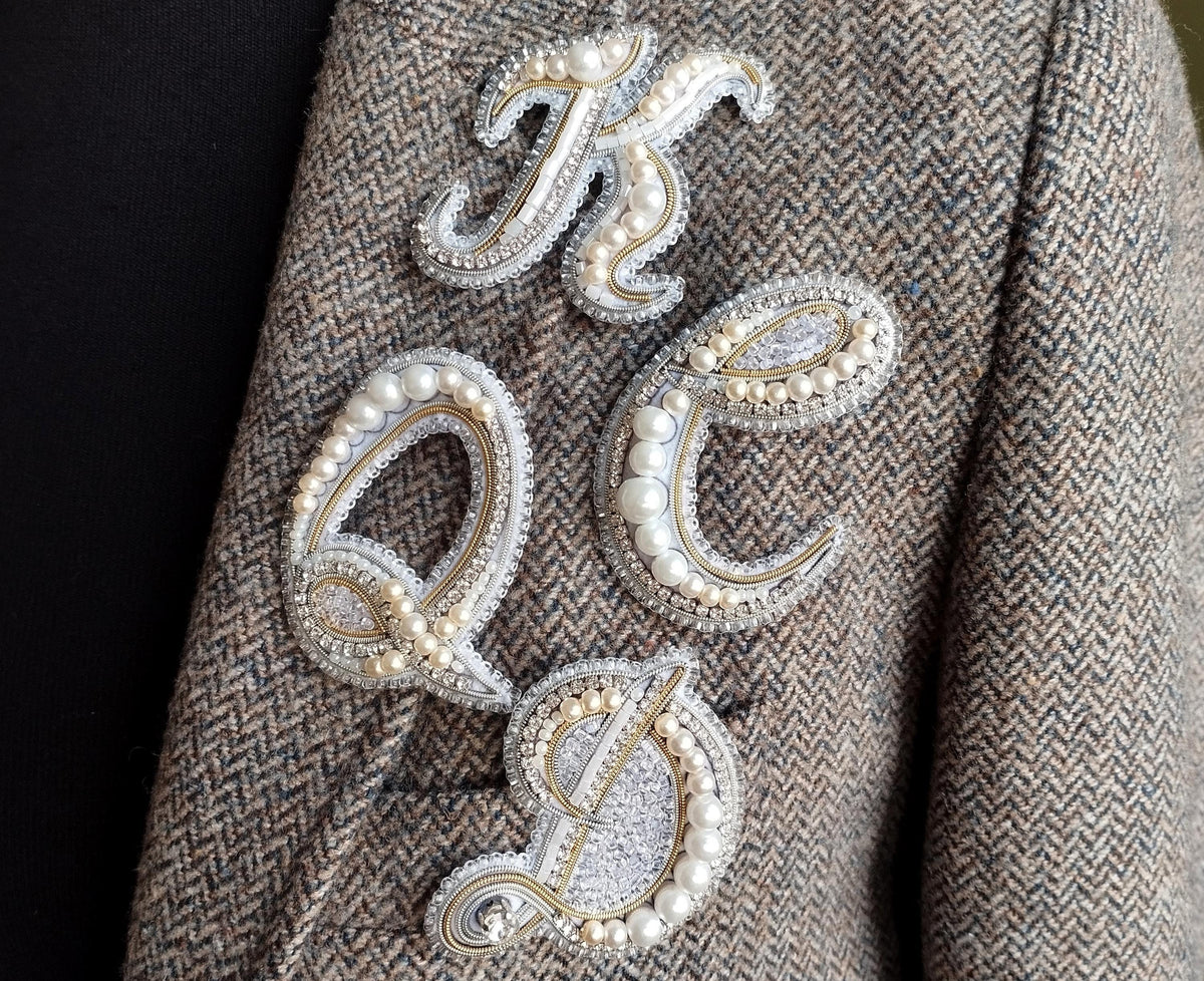 a close-up of a person's arm wearing a tweed jacket with a decorative embroidered letter on it.