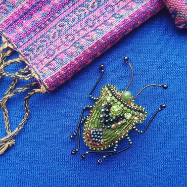 A colorful, intricately designed insect brooch is displayed on a blue fabric surface, with a pink and blue woven textile visible in the background.