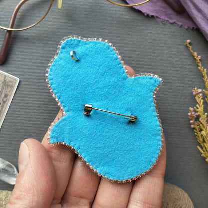 A hand holding a blue felt object with a pin attached to it, placed on a gray surface.