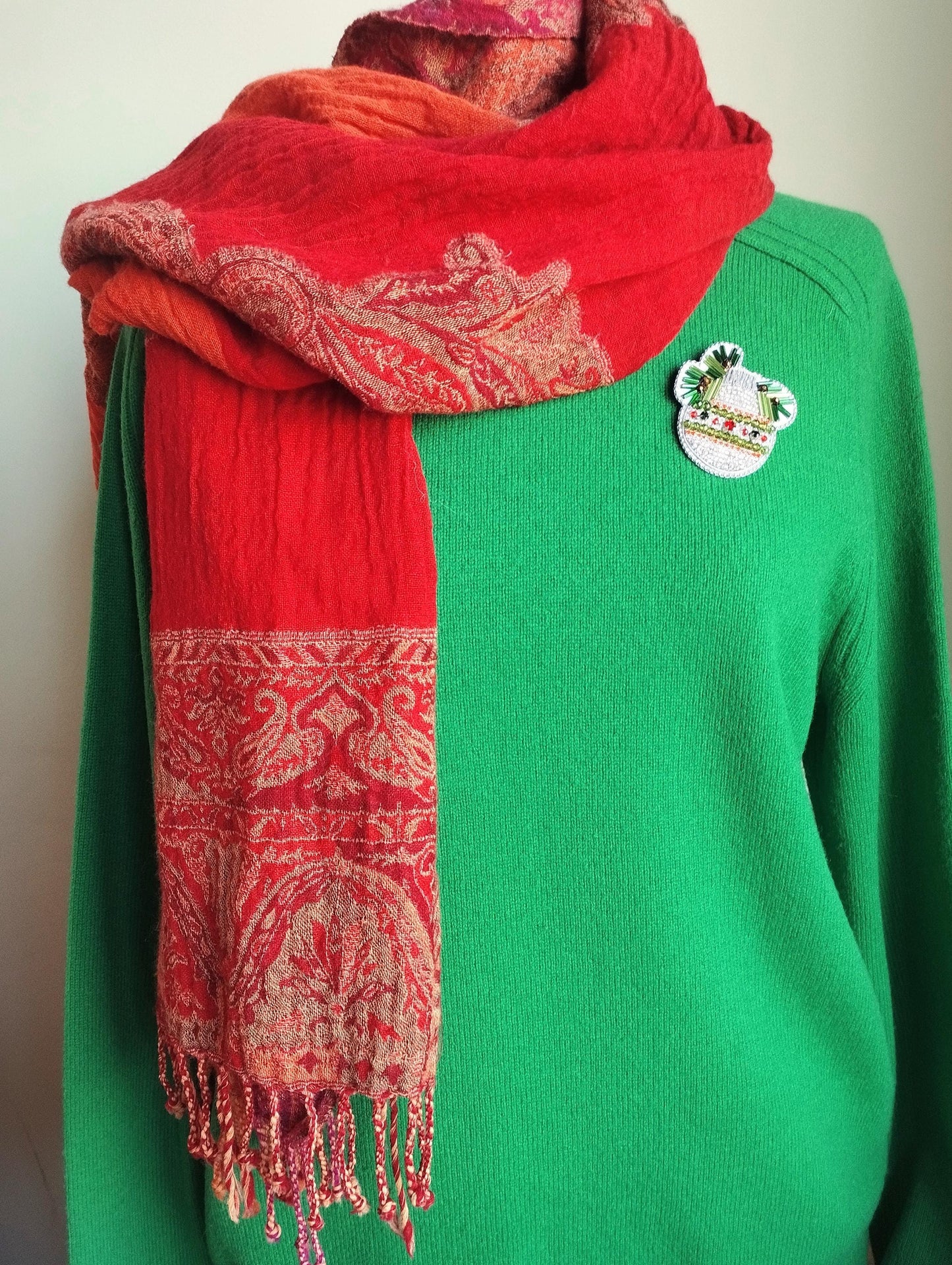 A mannequin is wearing a green sweater with a red and orange patterned scarf draped over it. The scarf has fringe details and a small pin on the sweater.