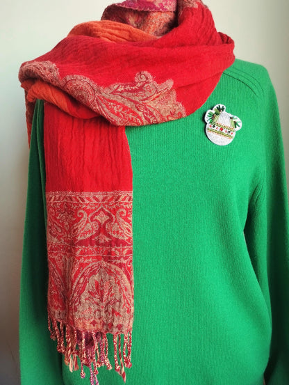 A mannequin is wearing a green sweater with a red and orange patterned scarf draped over it. The scarf has fringe details and a small pin on the sweater.