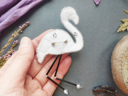 A hand holding a white felt flamingo pin with black legs and silver beads, surrounded by various craft materials and natural elements.