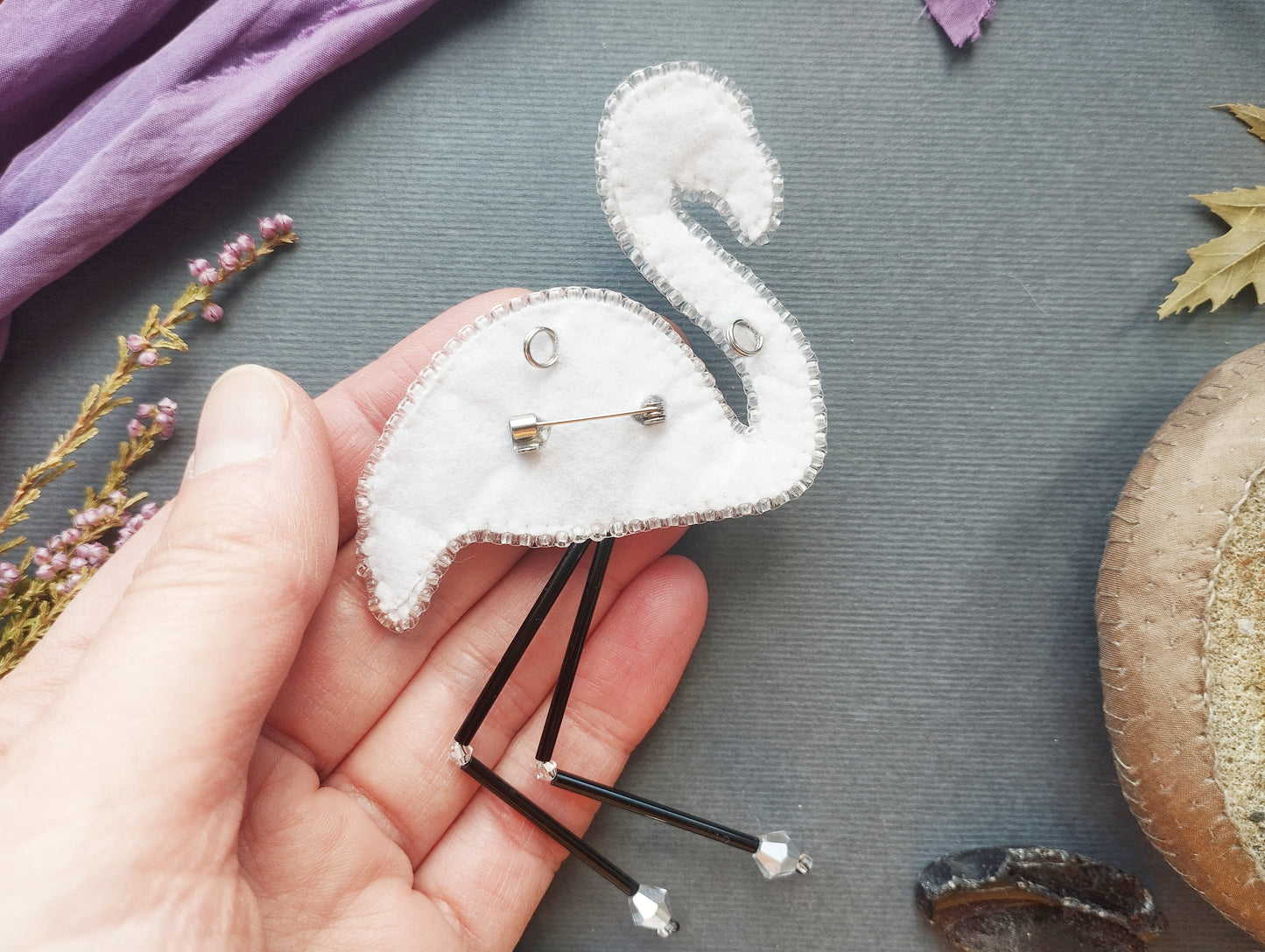 A hand holding a white felt flamingo pin with black legs and silver beads, surrounded by various craft materials and natural elements.